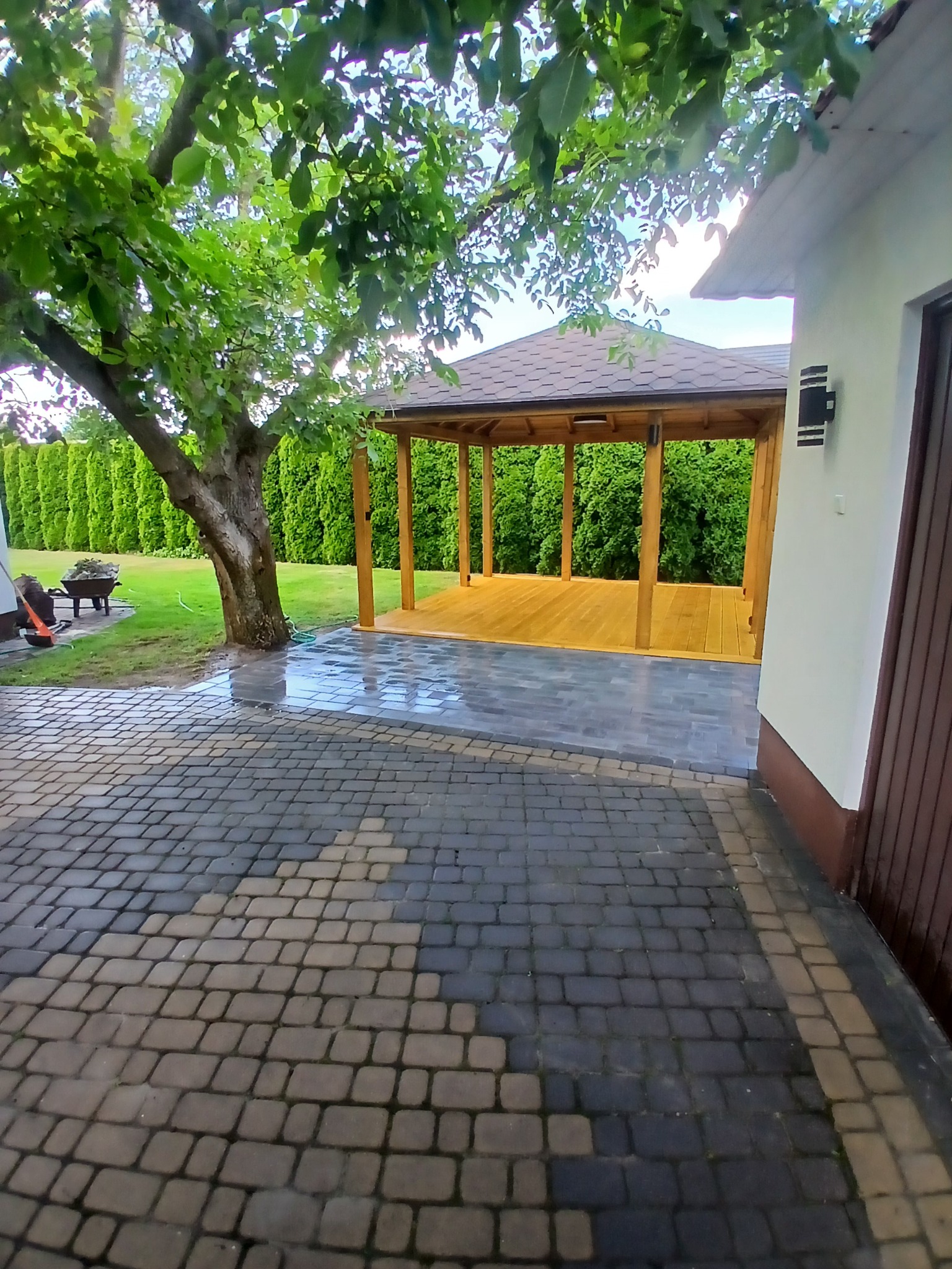 Drewniana altana z brązowym dachem, widoczna z perspektywy brukowanego podjazdu, otoczona zielenią drzew i krzewów. Elementy architektury ogrodowej w naturalnym otoczeniu.