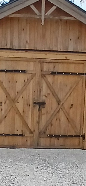 Drewniana altana z dwuskrzydłowymi drzwiami z metalowymi okuciami i zamkiem, widok z bliska na drzwi i fragment dachu, szara nawierzchnia przed budowlą.