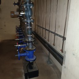 OLEKSANDR MAKSAI - Instalacja pionowych rur z zaworami, mocowanych do ściany za pomocą metalowych wsporników. Widoczne połączenia kołnierzowe i niebieskie zawory.