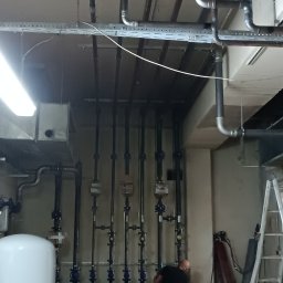 OLEKSANDR MAKSAI - Instalacja rur z zaworami w piwnicy, widoczny pracownik w trakcie montażu, butle z gazem i drabina. Rury prowadzone pod sufitem.