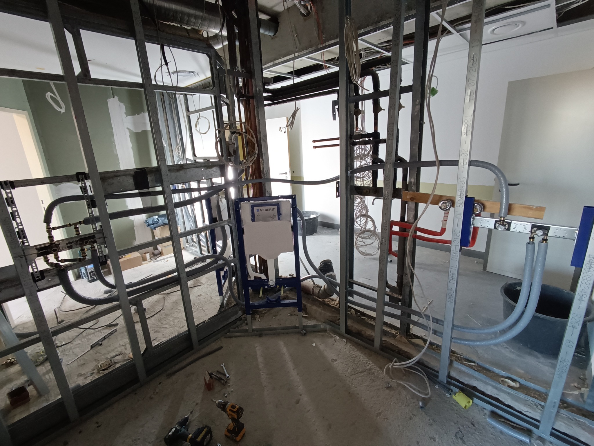 Instalacja podtynkowa Geberit Duofix w trakcie montażu, widoczne rury doprowadzające wodę, stelaż metalowy, narzędzia na podłodze. Surowy stan wykończenia wnętrza.
