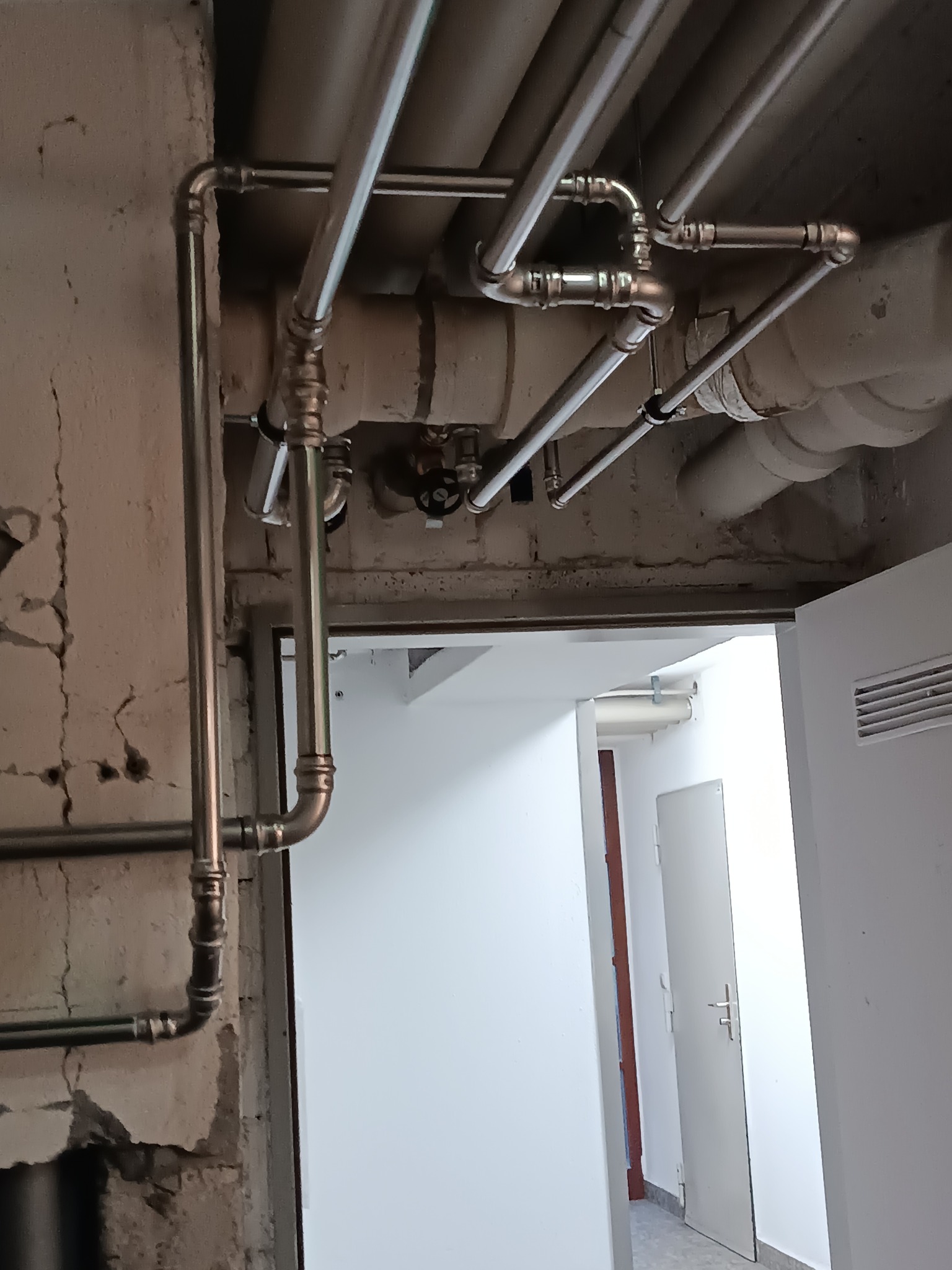 Instalacja hydrauliczna z rur chromowanych, widoczne połączenia i zawory, surowy beton na ścianie, w tle białe drzwi i korytarz. Nietypowe ułożenie rur.