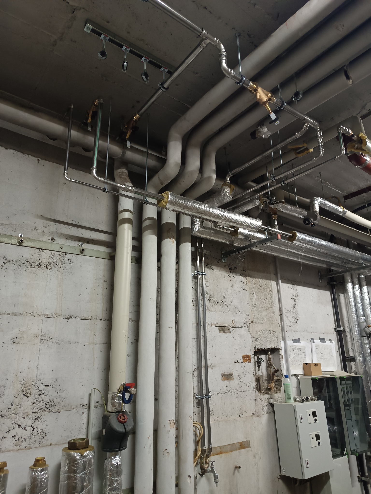 Kompleksowa instalacja rur w piwnicy z widocznymi zaworami, rurami izolowanymi i nieizolowanymi, oraz skrzynką kontrolną na ścianie z surowego betonu.