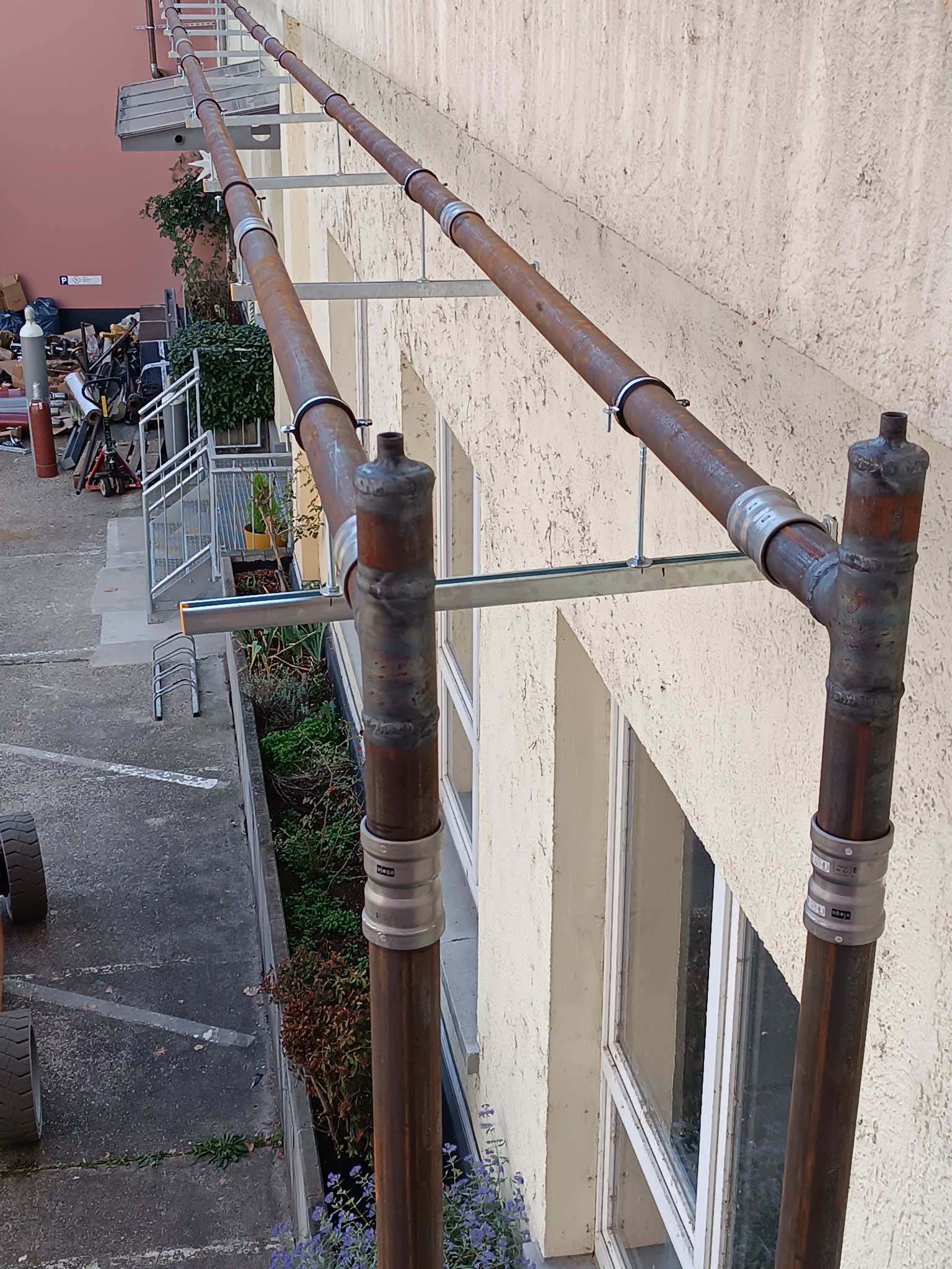 Zewnętrzna instalacja rurociągowa z brązowych rur, mocowana do ściany budynku za pomocą metalowych wsporników i obejm. Widoczne spawy i elementy montażowe.