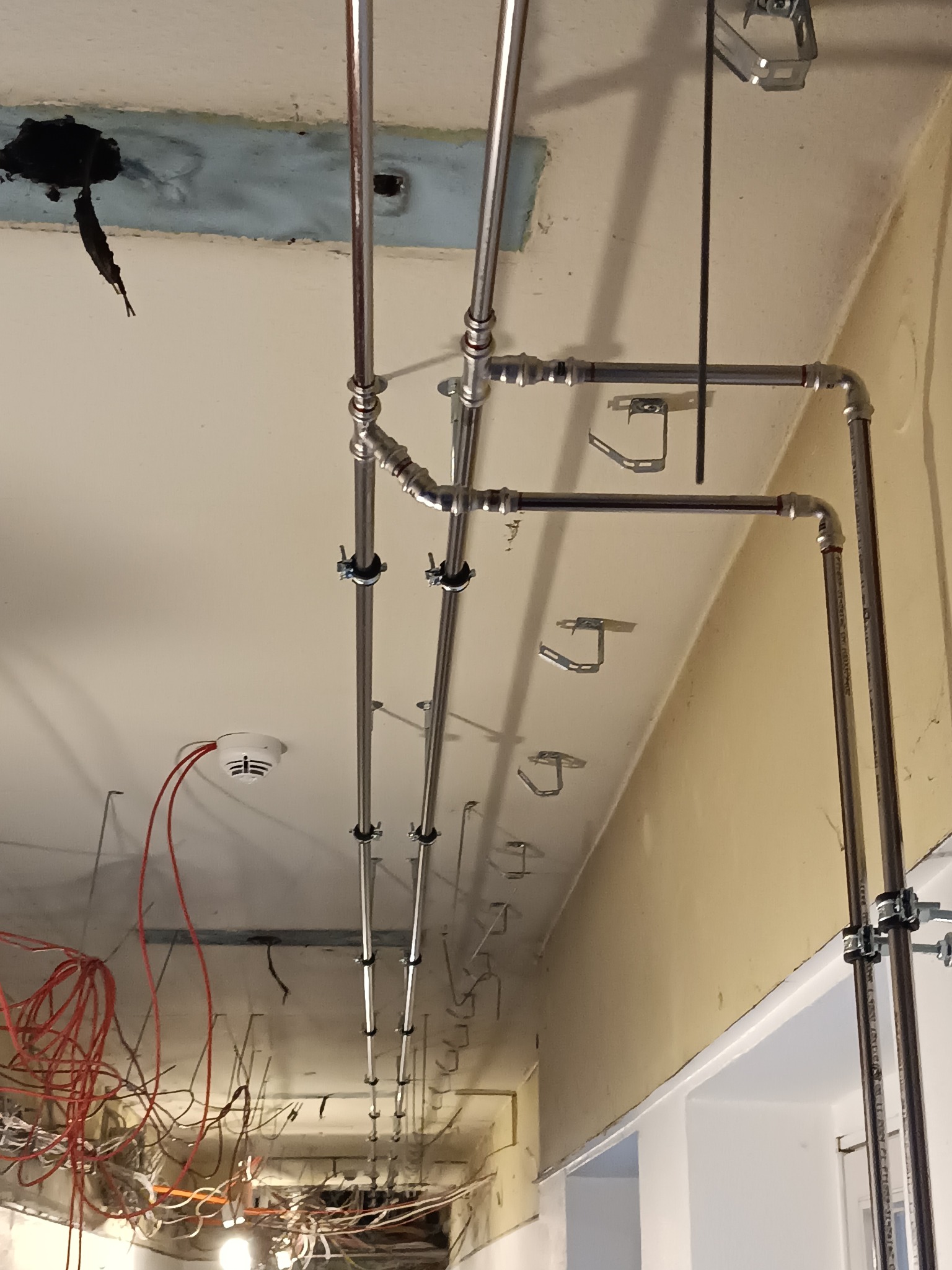 Montaż instalacji pod sufitem z widocznymi rurami, uchwytami i czujnikiem dymu. Widoczne kable i elementy konstrukcyjne sufitu podwieszanego.