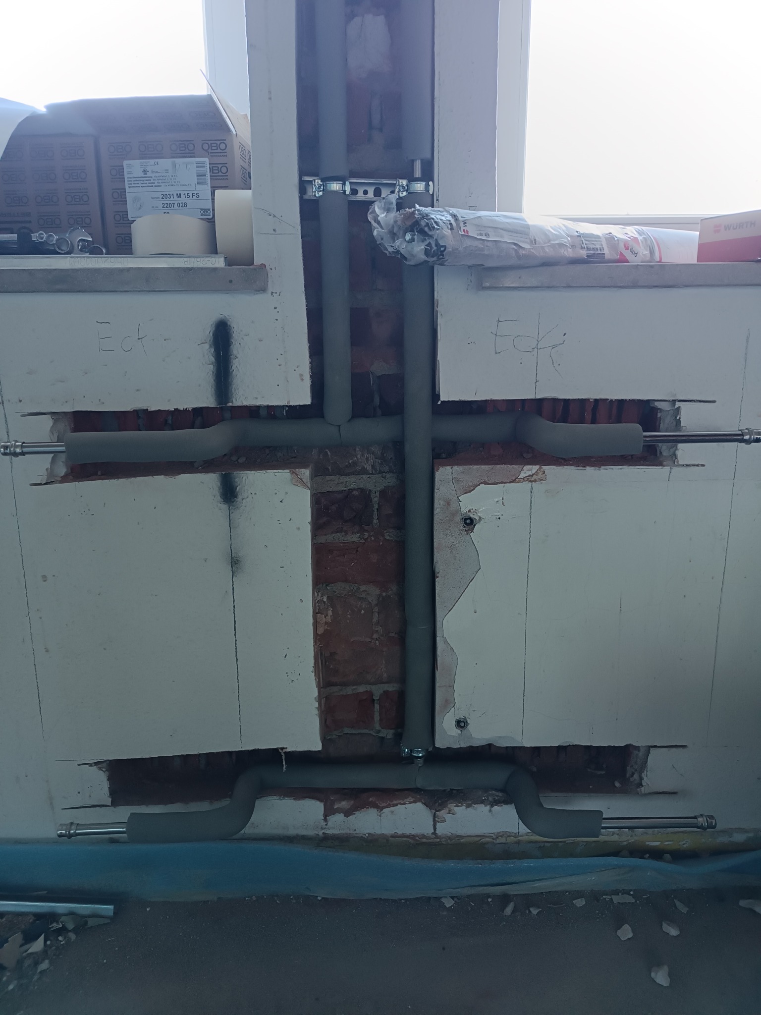 Szare rury instalacji grzewczej, częściowo zaizolowane, zamontowane w ścianie z cegły i płyt gipsowych. Widoczne elementy mocujące i fragment podłogi z folią ochronną.