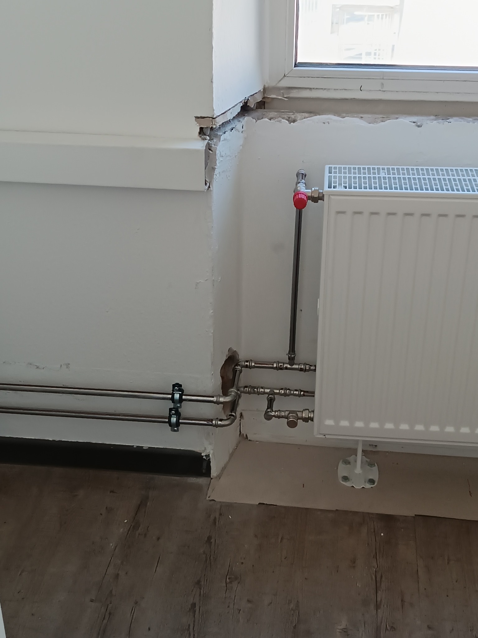 Biały grzejnik żeberkowy z instalacją w pomieszczeniu po remoncie w Warszawie. Widoczne rury i zawór termostatyczny. Podłoga drewnopodobna.