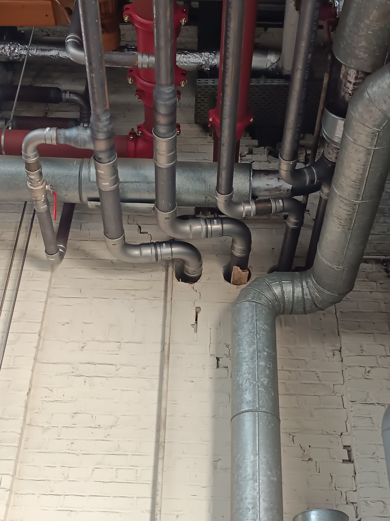 Skomplikowany układ rur grzewczych i hydraulicznych w piwnicy, z widocznymi połączeniami i zaworami, na tle białej, ceglanej ściany. Różne kolory rur wskazują na różne systemy.