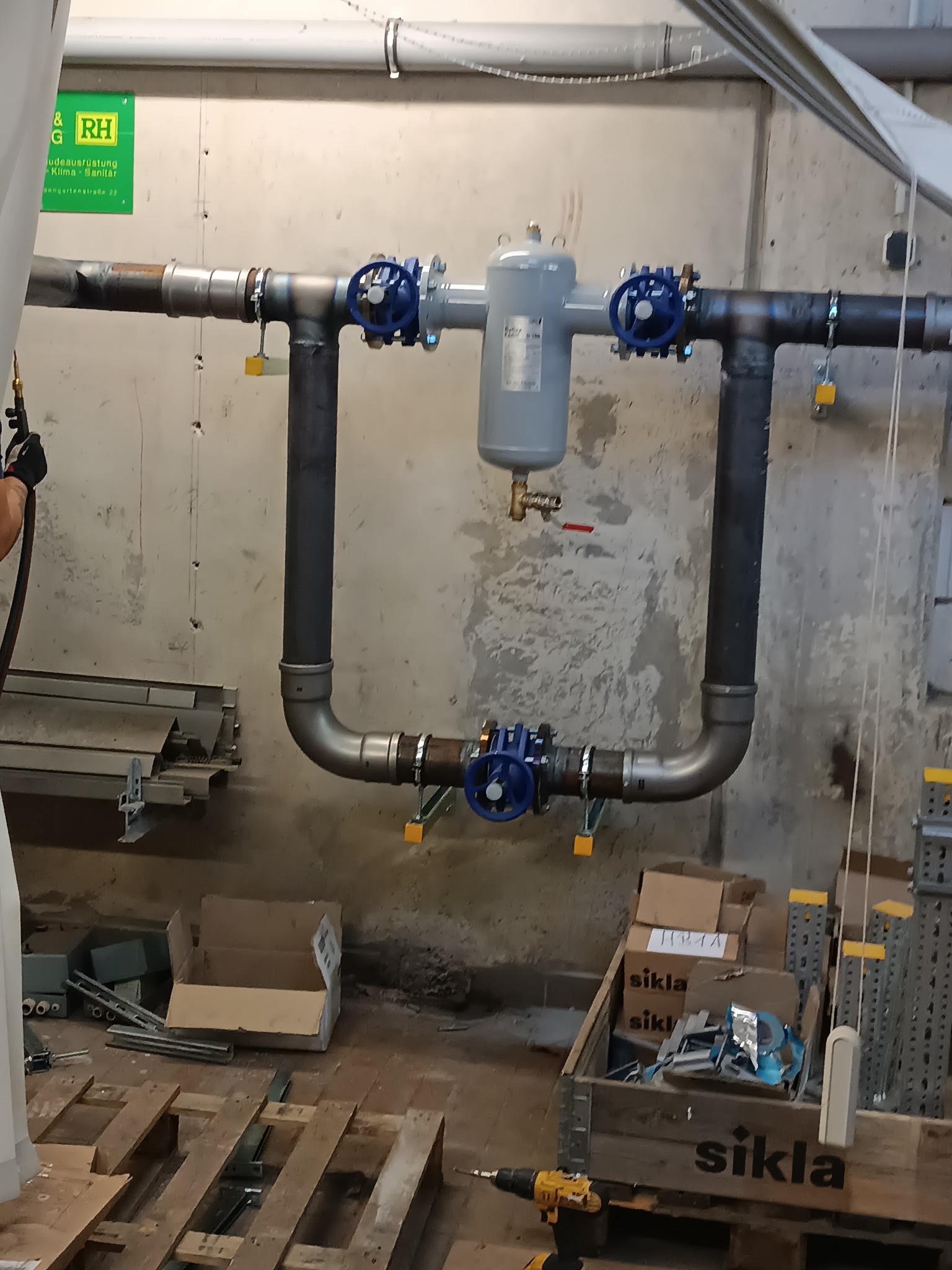 Zamknięty obieg instalacji z szarym zbiornikiem wyrównawczym i niebieskimi zaworami, zamontowany na szarej ścianie. Widoczne narzędzia i materiały montażowe.