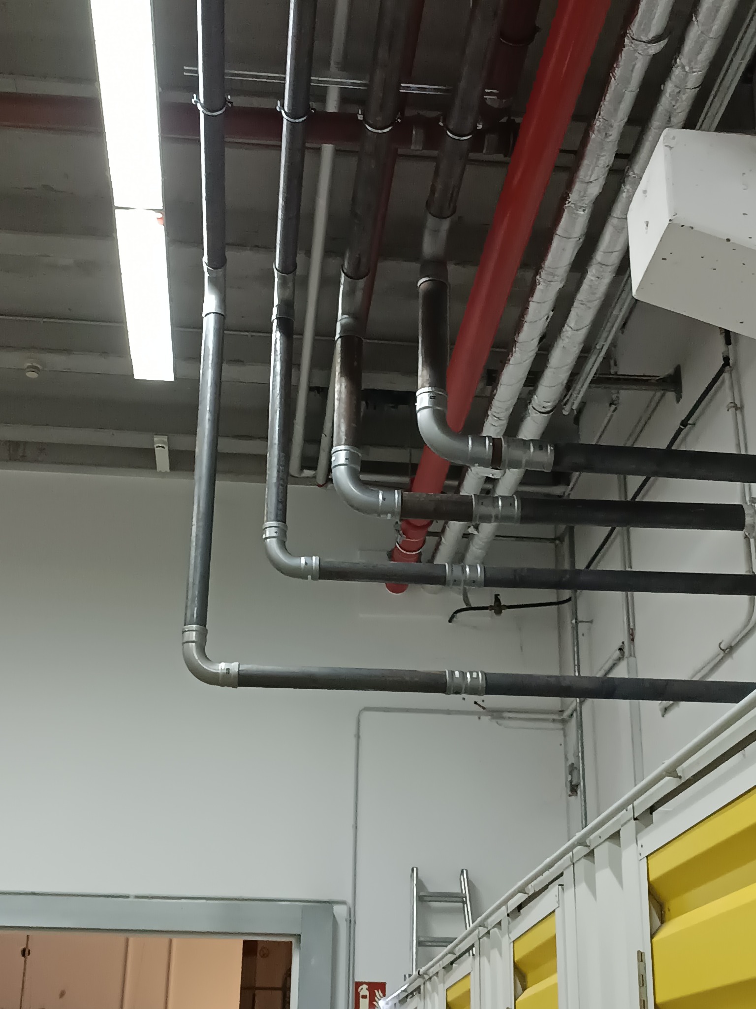 Różnokolorowe rury pod sufitem w hali, z widocznymi łączeniami i mocowaniami. W tle biała ściana i fragment żółtego kontenera.