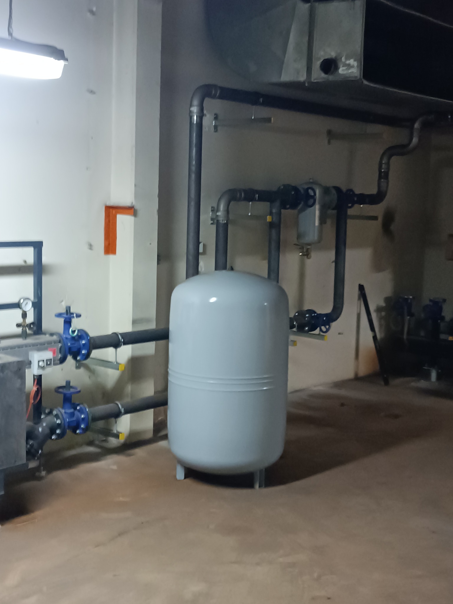 Szary zbiornik ciśnieniowy z niebieskimi zaworami i czarnymi rurami w kotłowni. Widoczne elementy instalacji grzewczej i hydraulicznej w pomieszczeniu technicznym.