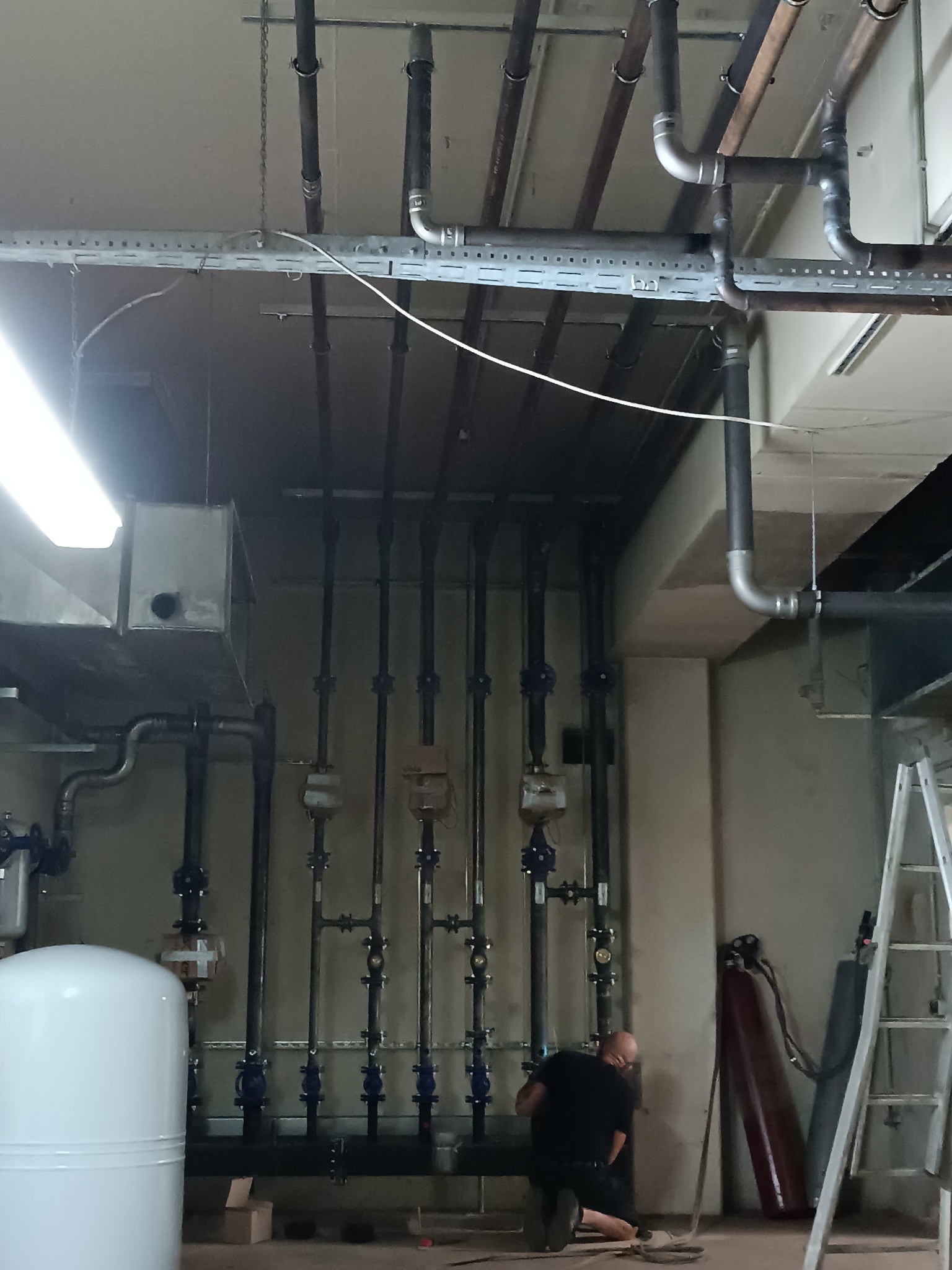 Instalacja rur z zaworami w piwnicy, widoczny pracownik w trakcie montażu, butle z gazem i drabina. Rury prowadzone pod sufitem.