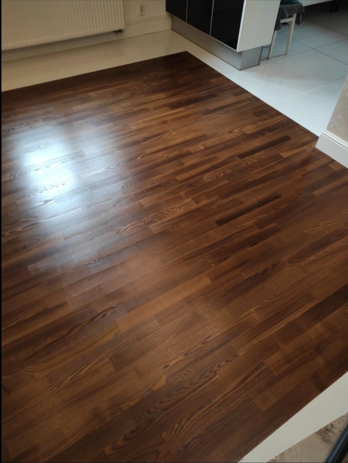 Ciemnobrązowa, lakierowana podłoga parkietowa z wyraźnym usłojeniem drewna, ułożona w pomieszczeniu z białymi listwami przypodłogowymi i fragmentem mebli.