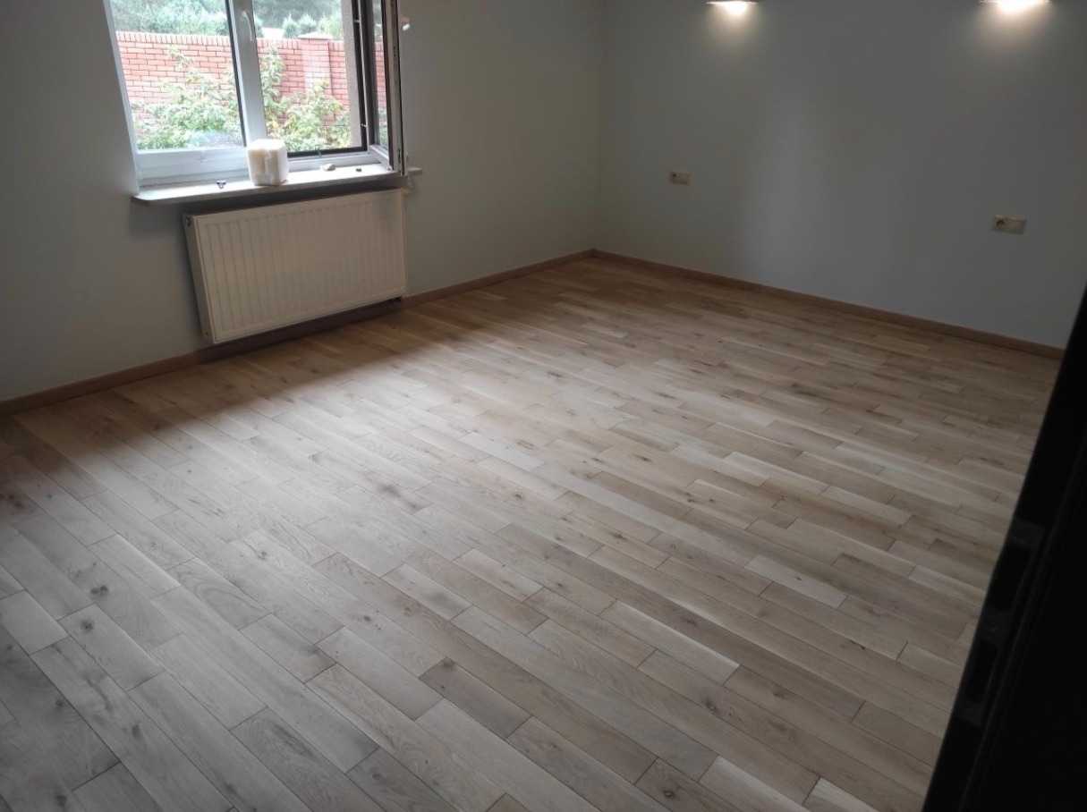 Jasny, odnowiony parkiet w pokoju z oknem i grzejnikiem. Świeżo położona podłoga z naturalnego drewna, widoczne listwy przypodłogowe i subtelne oświetlenie sufitowe.