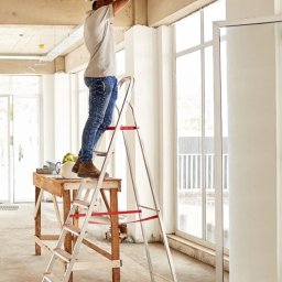 Bartłomiej Podlach - Elektryk w kasku na drabinie montuje korytko kablowe pod sufitem w jasnym pomieszczeniu. Widoczne narzędzia i elementy instalacji.