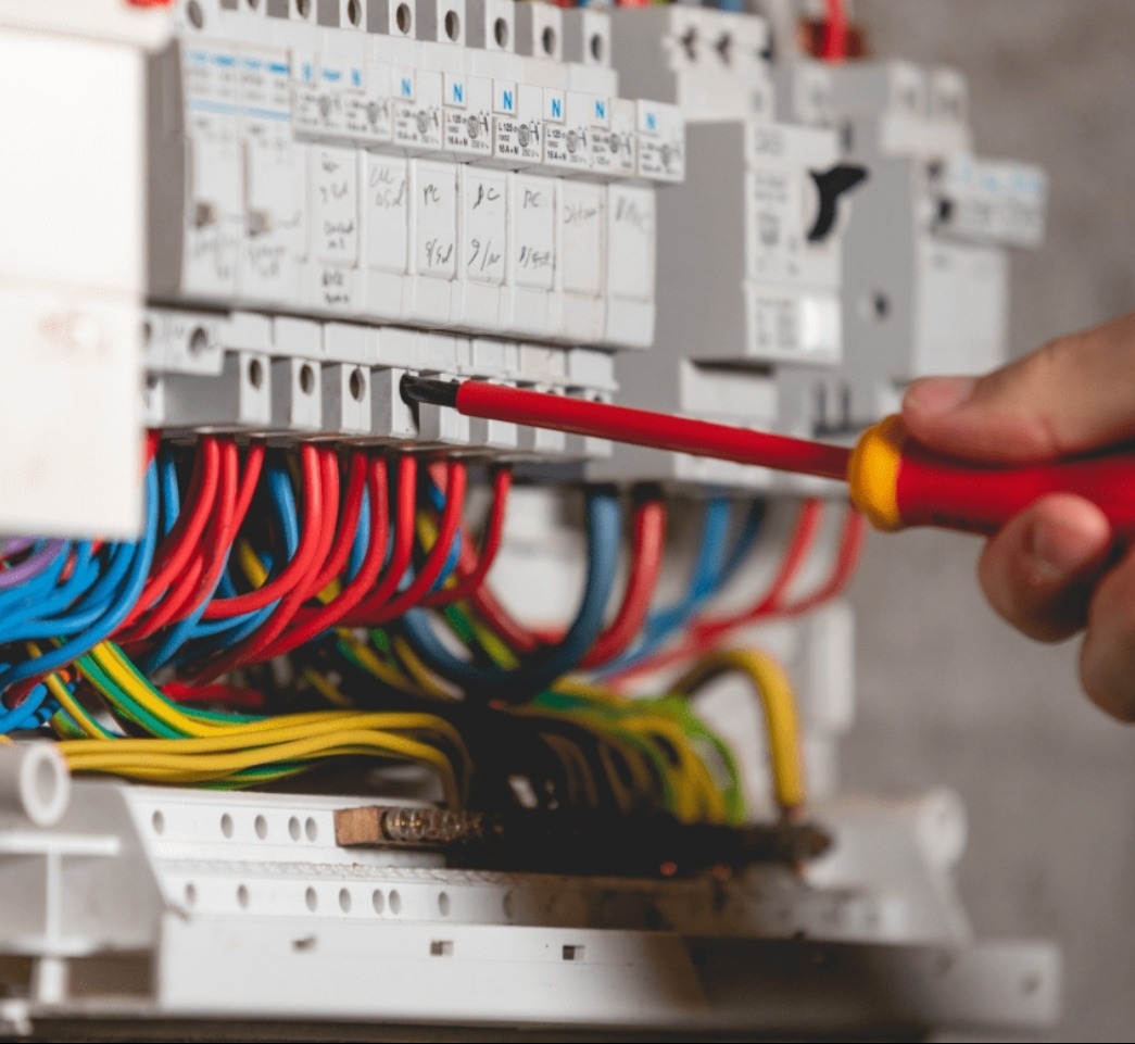 Elektryk w trakcie pracy przy rozdzielnicy, używając czerwonego śrubokręta. Widoczne kolorowe przewody i białe zabezpieczenia z opisami.