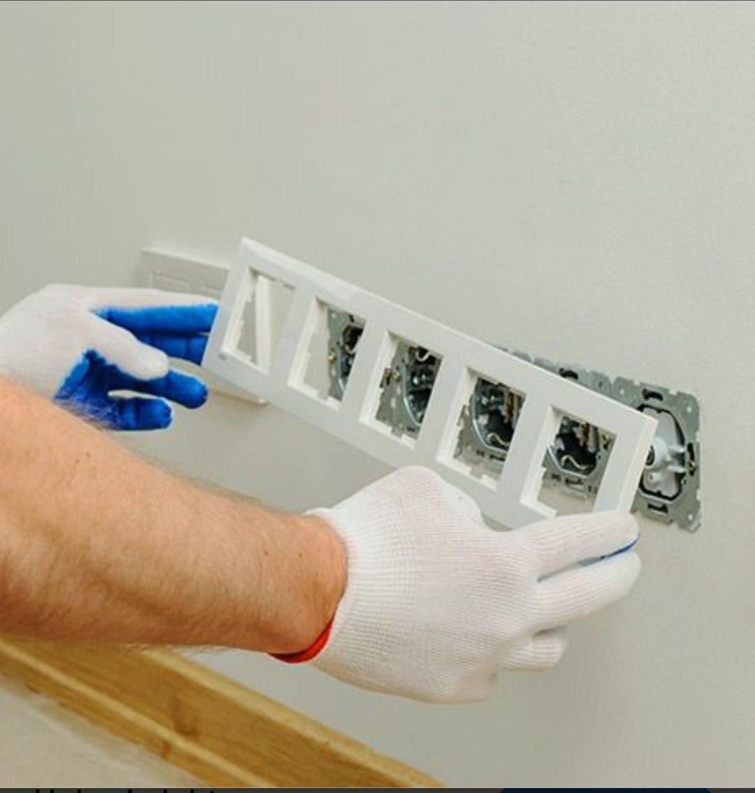 Elektryk w rękawicach montuje białą ramkę gniazdek elektrycznych na ścianie. Widoczne gniazdka i przewody. Precyzyjny montaż osprzętu elektrycznego.