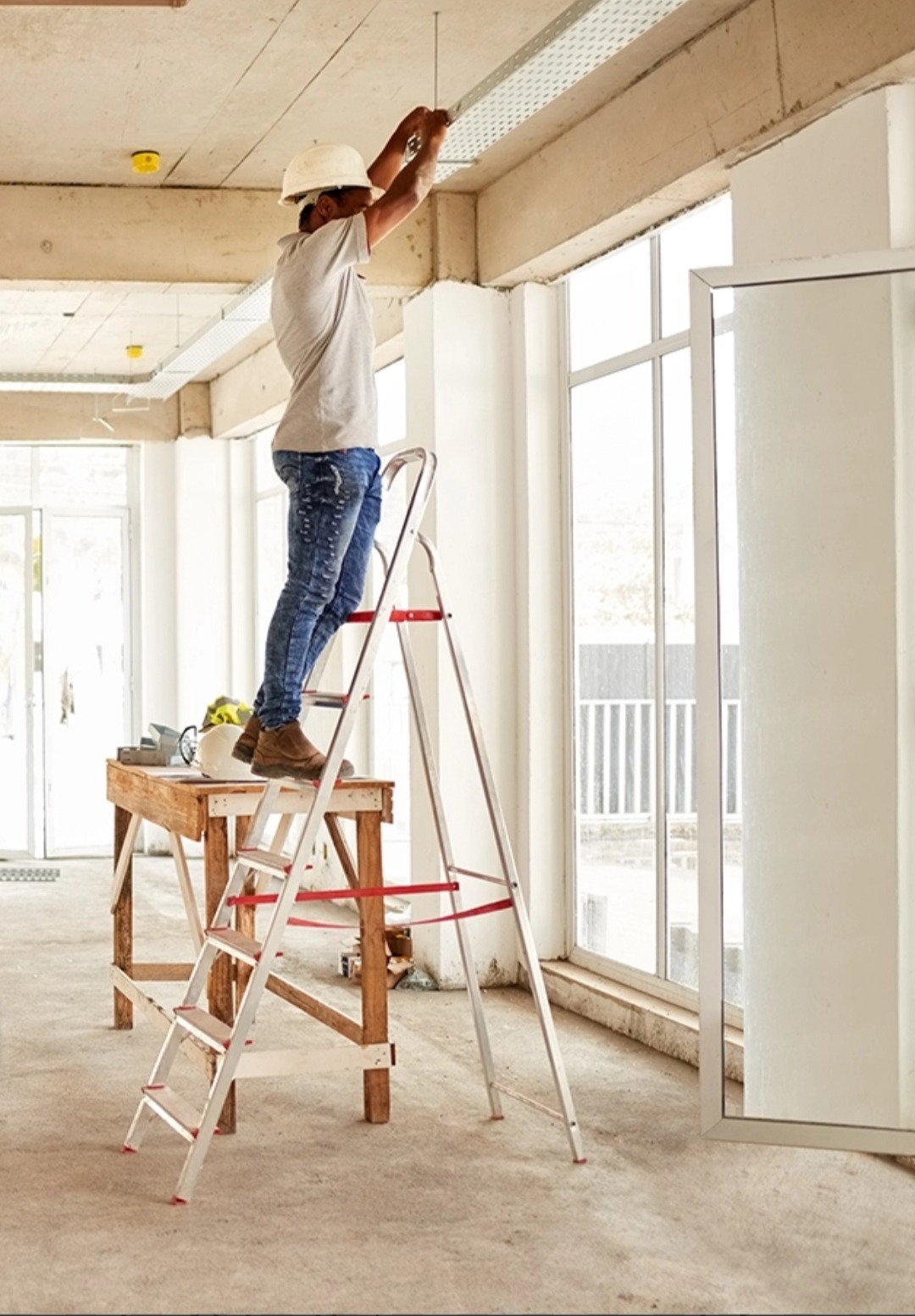 Elektryk w kasku na drabinie montuje korytko kablowe pod sufitem w jasnym pomieszczeniu. Widoczne narzędzia i elementy instalacji.