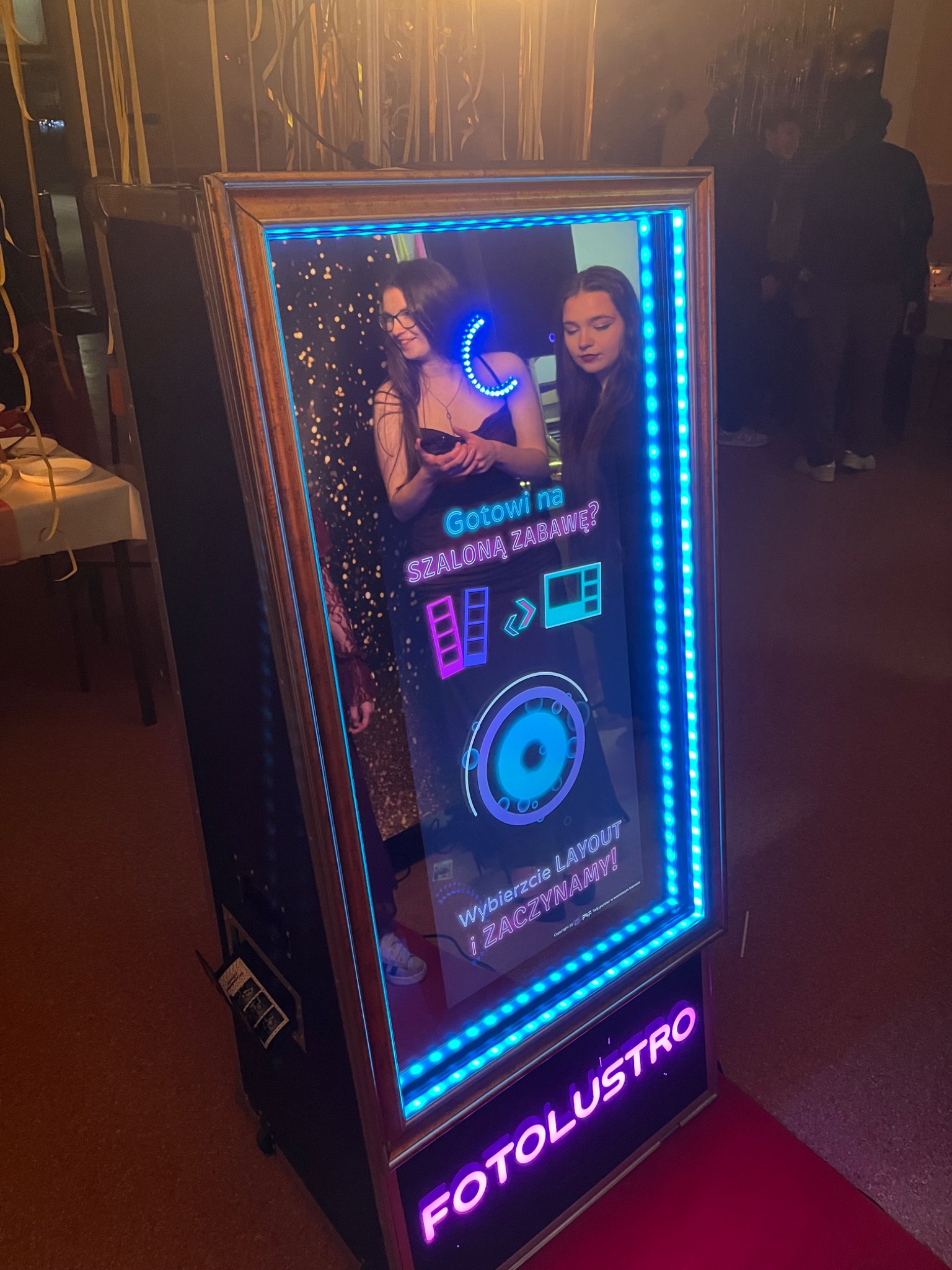 Fotolustro z niebieskim oświetleniem LED i odbijającymi się w lustrze osobami. Na ekranie interfejs wyboru layoutu. Widoczny napis 'FOTOLUSTRO' w kolorze fioletowym.