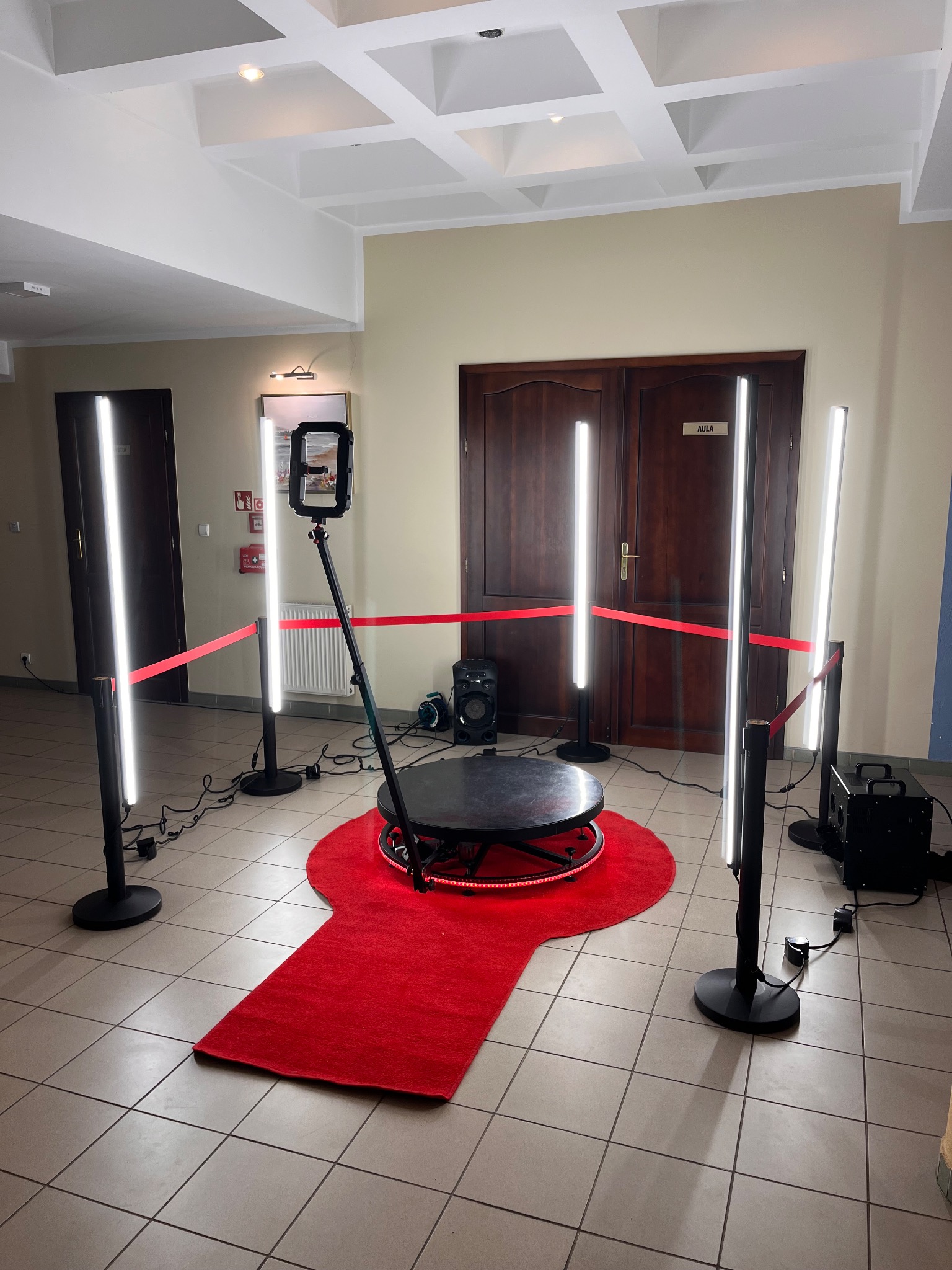 Kompletny zestaw fotobudki 360 z czerwonym dywanem, oświetleniem LED i barierkami, gotowy do użycia na imprezie w przestronnym wnętrzu.