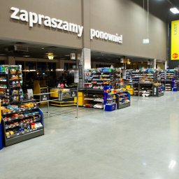 RENOVA SPÓŁKA Z OGRANICZONĄ ODPOWIEDZIALNOŚCIĄ - Jasna, lśniąca posadzka przemysłowa w supermarkecie, widok na rzędy kas z towarem. Czysta przestrzeń, żółto-czarne elementy wyposażenia, oświetlenie sufitowe.