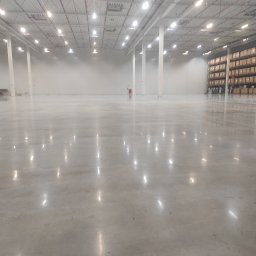 RENOVA SPÓŁKA Z OGRANICZONĄ ODPOWIEDZIALNOŚCIĄ - Błyszcząca, gładka posadzka przemysłowa w przestronnym magazynie z regałami w tle. Widoczne odbicia świateł na powierzchni podłogi.