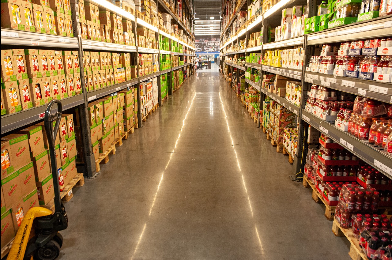 Błyszcząca, gładka posadzka przemysłowa w hali magazynowej z regałami pełnymi towarów. W tle widoczny wózek paletowy.