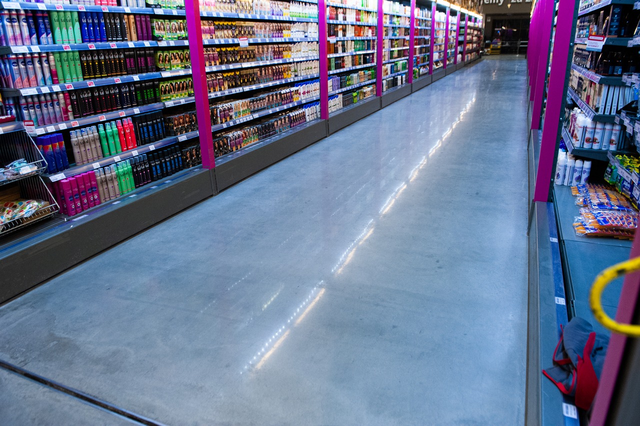 Błyszcząca, szara posadzka przemysłowa w supermarkecie, odbijająca światło. Półki z towarem po obu stronach alejki. Widok w perspektywie, lekko z góry.