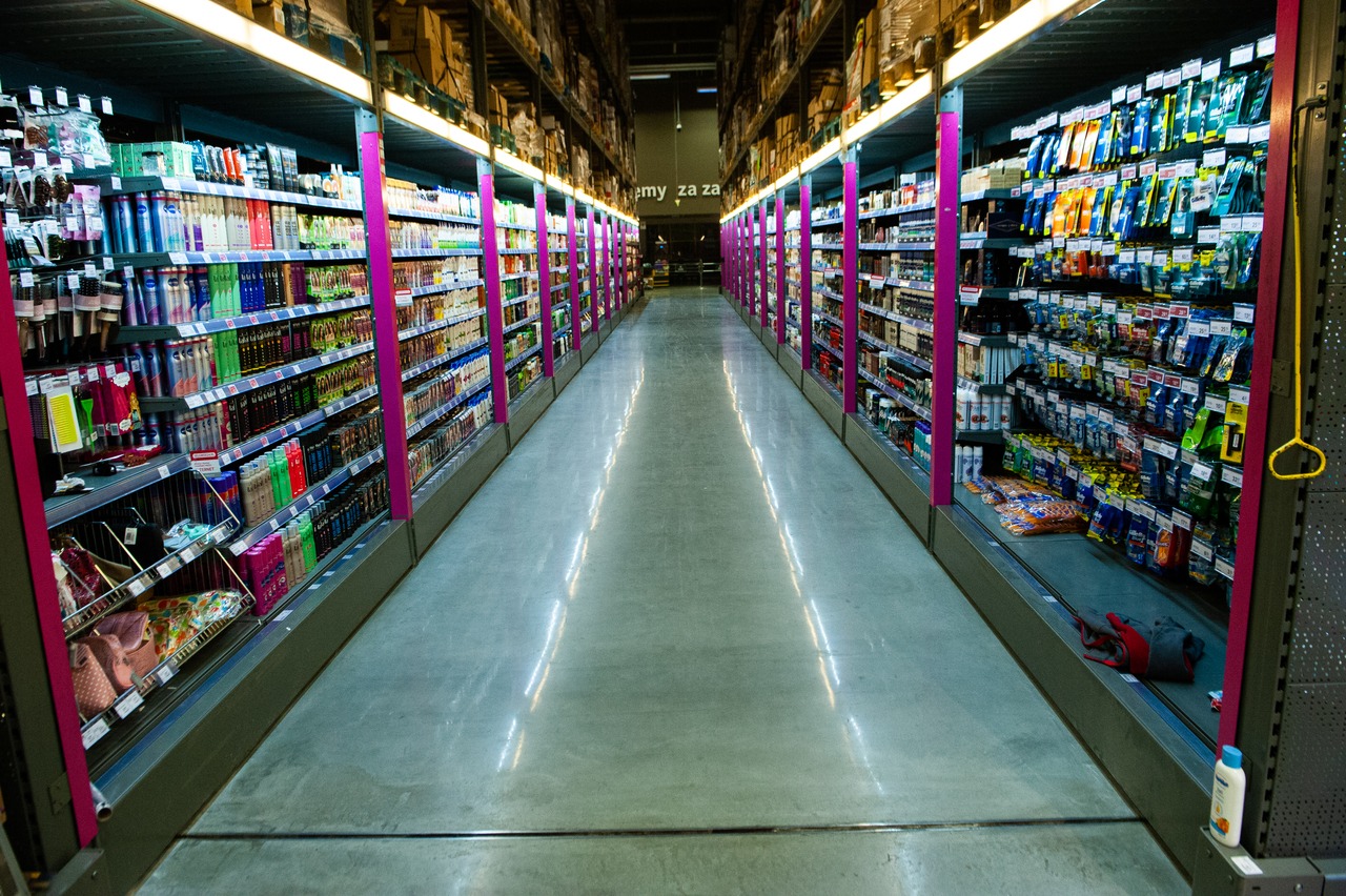 Długa perspektywa hali magazynowej z równymi rzędami regałów wypełnionych towarem, oświetlona jasnym światłem, z szarą, gładką posadzką przemysłową.