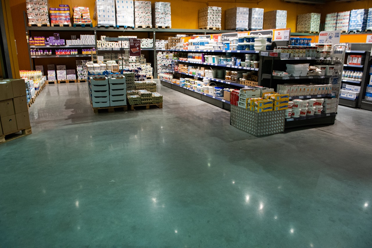 Błyszcząca, szara posadzka przemysłowa w hali magazynowej z regałami pełnymi towarów. Widoczne refleksy świetlne na gładkiej powierzchni. Perspektywa z niskiego kąta.