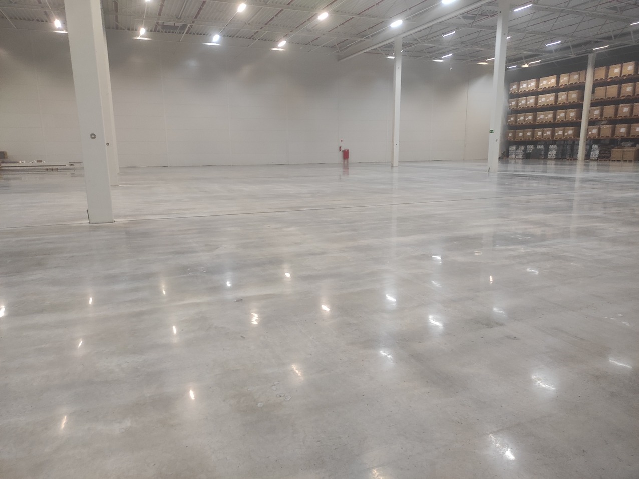 Błyszcząca, szara posadzka przemysłowa w przestronnym magazynie z regałami w tle. Widoczne odbicia światła od lamp sufitowych na gładkiej powierzchni.