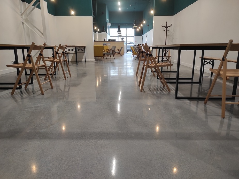 Błyszcząca, szara posadzka przemysłowa w przestronnym biurze z drewnianymi krzesłami i stołami. Widoczne odbicia światła na gładkiej powierzchni. Nowoczesne wnętrze.