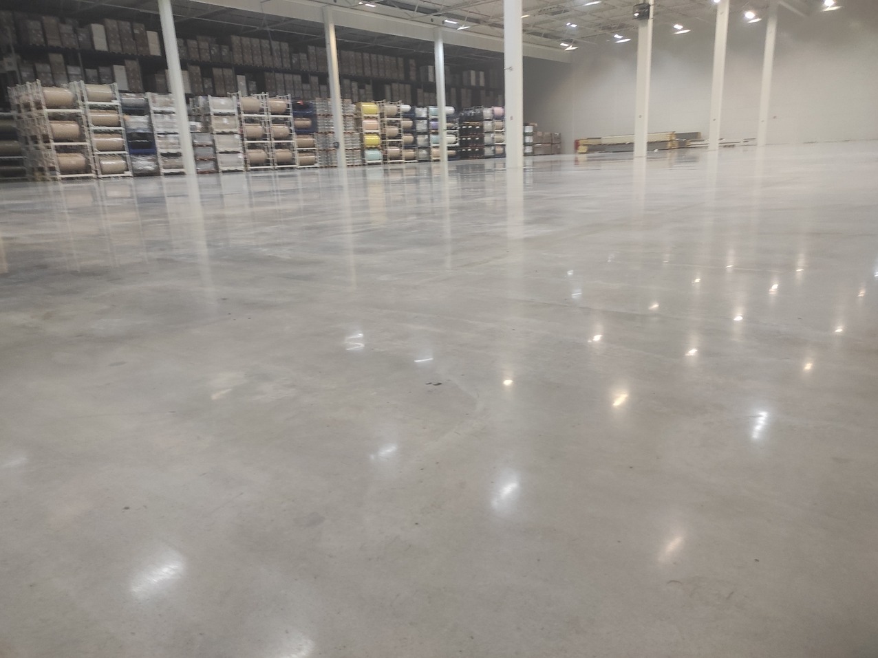 Błyszcząca, szara posadzka przemysłowa w magazynie z regałami w tle. Widoczne odbicia świateł w posadzce. Minimalistyczny kadr, podkreślający gładkość i równość powierzchni.