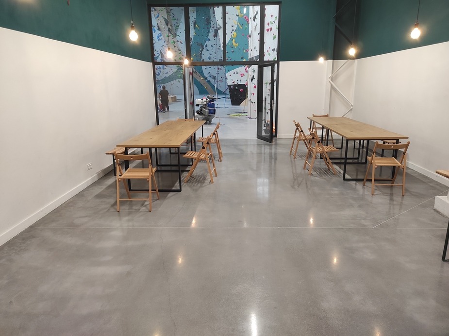 Błyszcząca, szara posadzka przemysłowa w przestronnym wnętrzu z drewnianymi stołami i krzesłami. W tle widoczna ściana wspinaczkowa i maszyna czyszcząca.