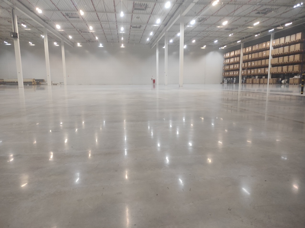 Błyszcząca, gładka posadzka przemysłowa w przestronnym magazynie z regałami w tle. Widoczne odbicia świateł na powierzchni podłogi.