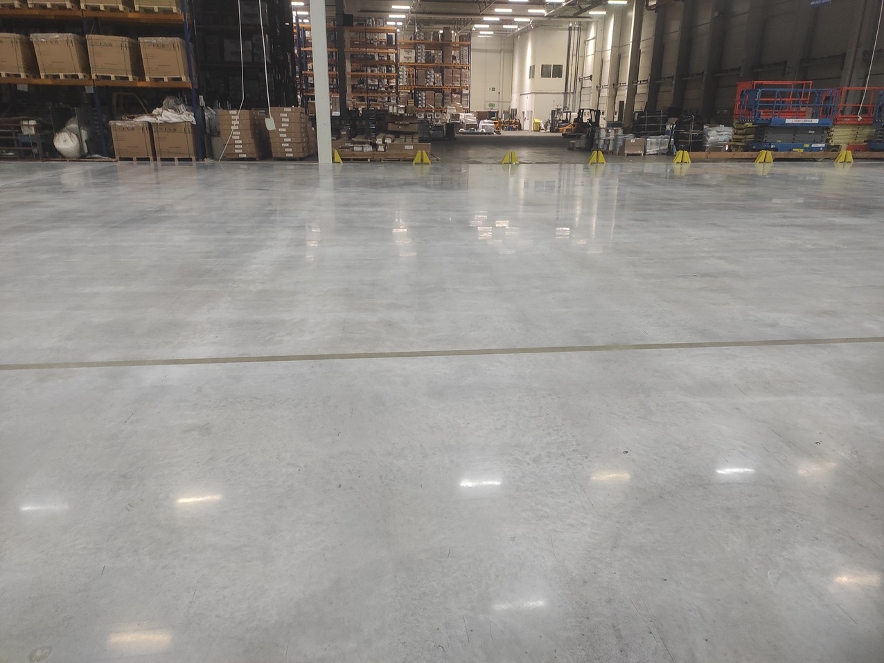 Błyszcząca, szara posadzka przemysłowa w hali magazynowej z widocznymi regałami i wózkiem widłowym w tle. Delikatna linia podziału na posadzce.