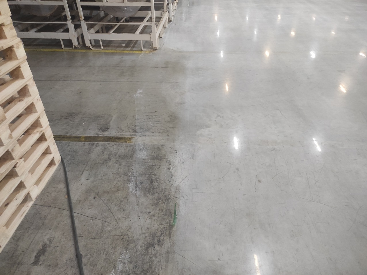 Częściowo wyczyszczona, betonowa posadzka przemysłowa z widocznymi refleksami światła, paletami i metalowymi konstrukcjami w tle. Widoczna różnica w czystości.