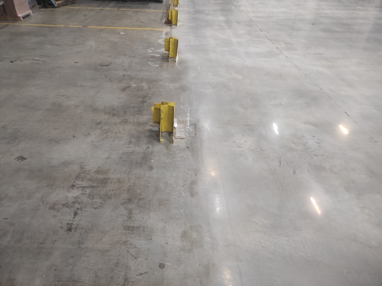 Szara, gładka posadzka przemysłowa z żółtymi odbojnikami w hali magazynowej. Widoczne ślady użytkowania i refleksy świetlne na powierzchni.