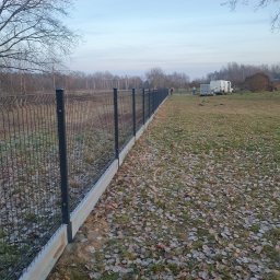 USŁUGI SPAWALNICZE KRYSTIAN PAWŁOWSKI - Długi, panelowy płot z podmurówką, ciągnący się wzdłuż działki pokrytej liśćmi. W tle widoczny las i ekipa montażowa z samochodem dostawczym.