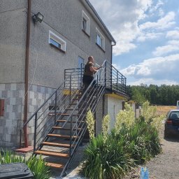 USŁUGI SPAWALNICZE KRYSTIAN PAWŁOWSKI - Metalowe schody z drewnianymi stopniami prowadzą na balkon budynku. Mężczyzna czyści balustradę. W tle zaparkowane auto i bus. Na pierwszym planie kwitnące juki.