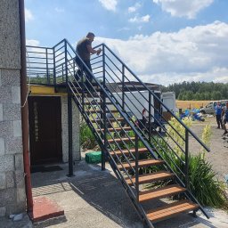 USŁUGI SPAWALNICZE KRYSTIAN PAWŁOWSKI - Zewnętrzne schody metalowe z drewnianymi stopniami, malowane przez mężczyznę. W tle budynek, pojazdy i osoby. Dzień słoneczny, błękitne niebo z chmurami.