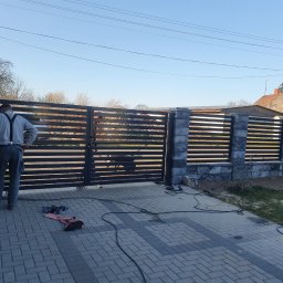 USŁUGI SPAWALNICZE KRYSTIAN PAWŁOWSKI - Nowoczesna brama wjazdowa z poziomych profili, częściowo zamontowana. Widoczny monter w szelkach i narzędzia na kostce brukowej. Słupki z szarego kamienia.