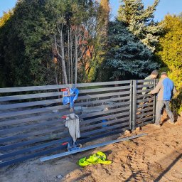 USŁUGI SPAWALNICZE KRYSTIAN PAWŁOWSKI - Montaż przęsła ogrodzeniowego z poziomych profili stalowych przez dwóch monterów. Widoczne narzędzia i odzież robocza. W tle zieleń drzew i budynek.