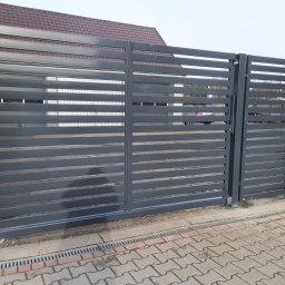 USŁUGI SPAWALNICZE KRYSTIAN PAWŁOWSKI - Nowoczesna, przesuwna brama wjazdowa z poziomych, szarych profili, zamontowana między dwoma kamiennymi słupkami. Widoczny fragment posesji i kostka brukowa.