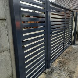 USŁUGI SPAWALNICZE KRYSTIAN PAWŁOWSKI - Nowoczesna, pozioma brama wjazdowa z metalowymi lamelami w kolorze grafitowym, zamontowana przy betonowym słupku. Widoczny zamek i uchwyt.