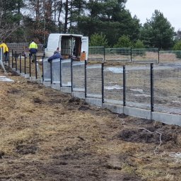 USŁUGI SPAWALNICZE KRYSTIAN PAWŁOWSKI - Montaż ogrodzenia panelowego na betonowej podmurówce. Widoczni pracownicy w odblaskowych ubraniach i biały bus w tle. Teren trawiasty, częściowo rozkopany.