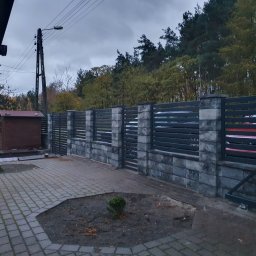 USŁUGI SPAWALNICZE KRYSTIAN PAWŁOWSKI - Nowoczesne ogrodzenie z szarego kamienia i metalowych paneli, widok z zewnątrz posesji, brama wjazdowa częściowo otwarta, jesienne otoczenie.