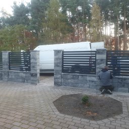 USŁUGI SPAWALNICZE KRYSTIAN PAWŁOWSKI - Nowoczesne ogrodzenie z szarych bloczków i poziomych, czarnych przęseł, z białym busem w tle i osobą montującą elementy. Widok z zewnątrz posesji.