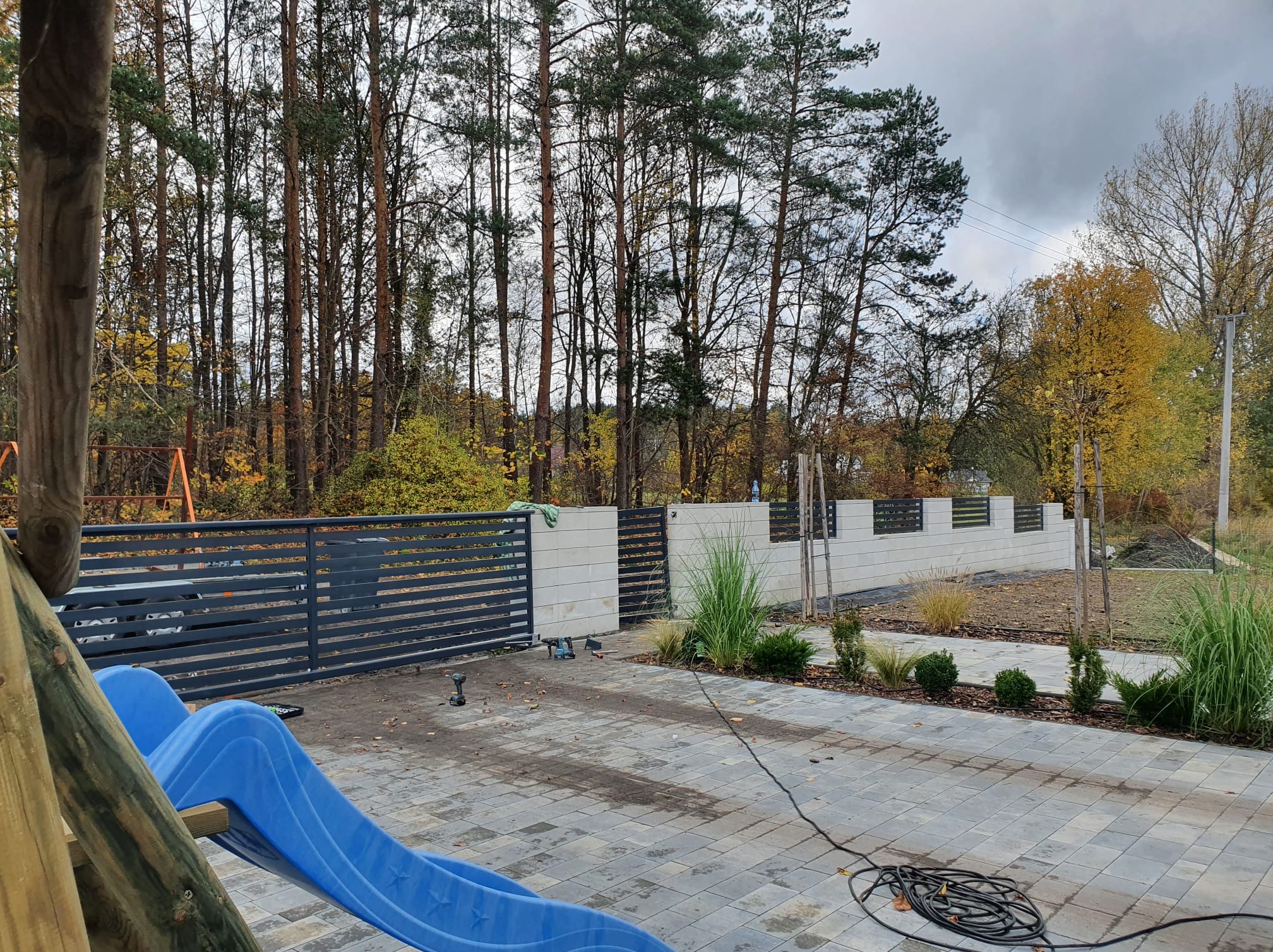 Nowoczesne ogrodzenie panelowe w kolorze grafitowym, z elementami murowanymi, otoczone zielenią i kostką brukową. Widoczne narzędzia montażowe.