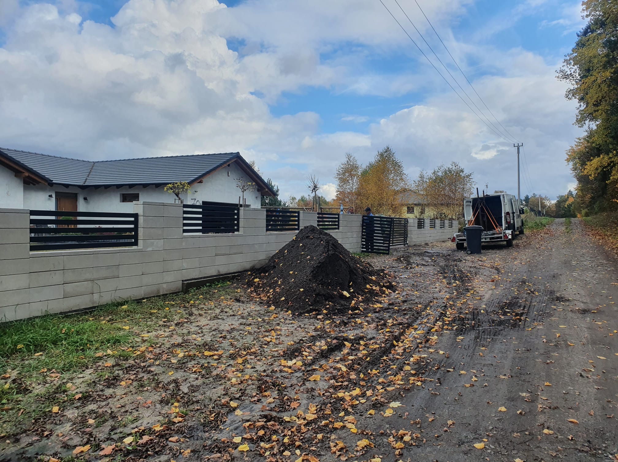 Nowoczesne ogrodzenie z betonowych bloczków i poziomych, czarnych przęseł metalowych, widoczne w trakcie montażu, z hałdą ziemi i samochodem dostawczym w tle.
